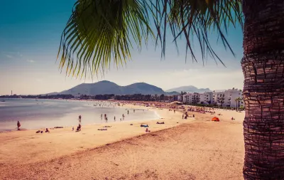 Crowded Mediterranean beach with mountains, palm tree, white apartments, and clear blue sky